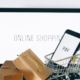 small cardboard boxes in a pile beside a small shopping cart with a phone inside it, atop a laptop